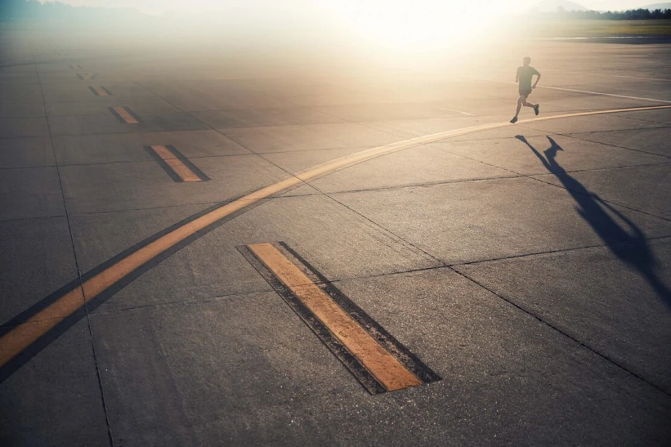Adult male running outdoors on concrete surface with yellow markings in bright sunlight. Shadow visible. Distant mountains.