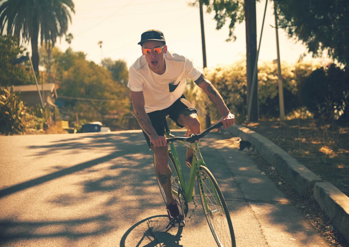 Adult male rides a green fixed-gear bicycle on a paved road during the daytime, with houses and trees visible in the background.