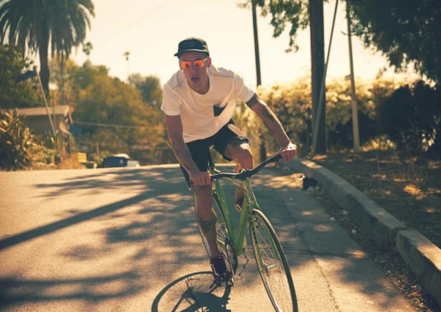 Adult male rides a green fixed-gear bicycle on a paved road during the daytime, with houses and trees visible in the background.