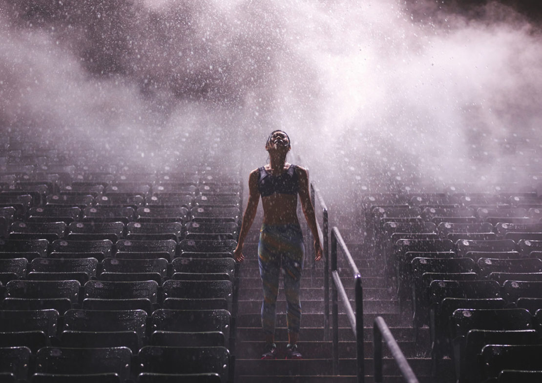 Athletic woman stands in an outdoor stadium, looking up into falling rain. Backlit by stadium lights, rows of empty seating visible.