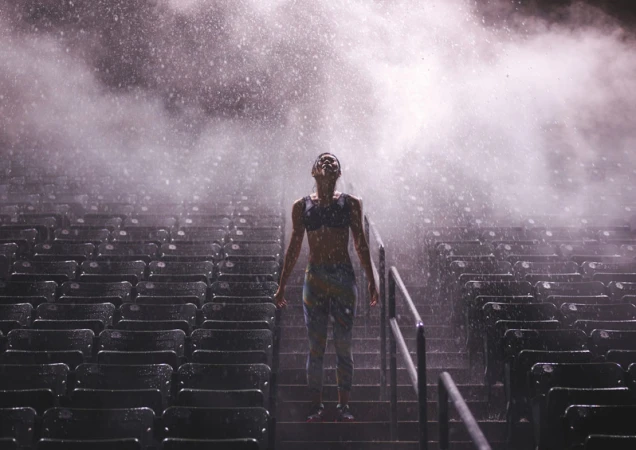 Athletic woman stands in an outdoor stadium, looking up into falling rain. Backlit by stadium lights, rows of empty seating visible.