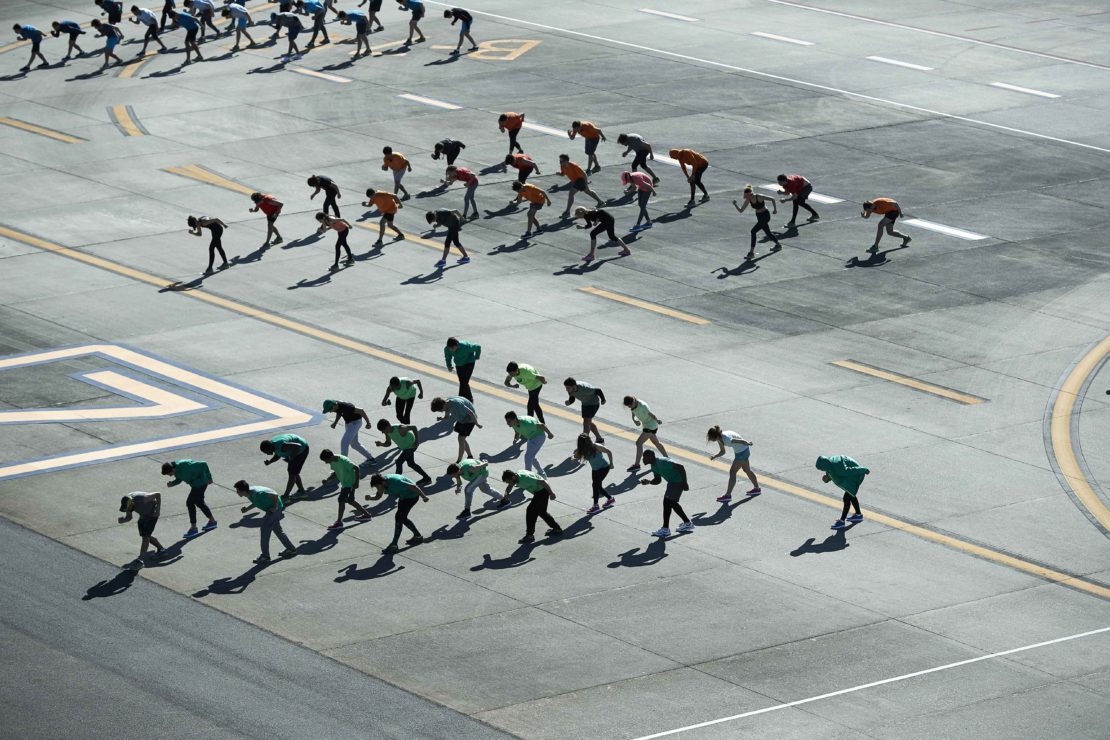 Aerial view of a large group of adults bending over on a gray concrete surface with yellow lines and shadows on a sunny day.
