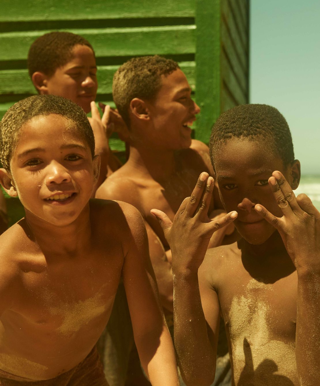 Group of smiling African boys gathered outdoors with playful hand gestures, showcasing a beachy, sunny scene with a green backdrop.