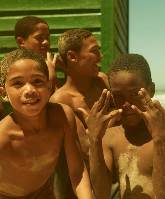 Group of smiling African boys gathered outdoors with playful hand gestures, showcasing a beachy, sunny scene with a green backdrop.
