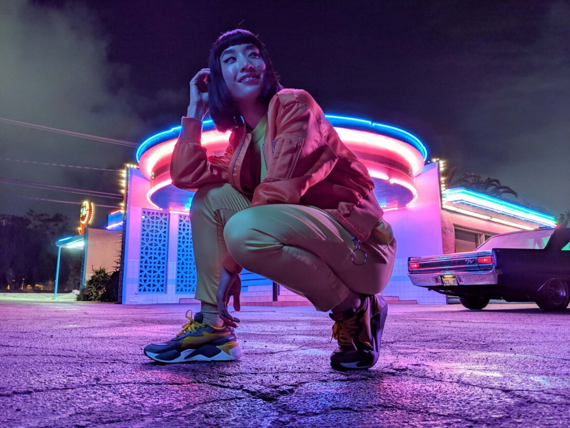 Woman in orange jacket and yellow pants crouching in front of a retro-style building with neon lights at dusk or night.