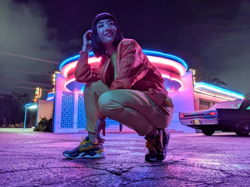 Woman in orange jacket and yellow pants crouching in front of a retro-style building with neon lights at dusk or night.