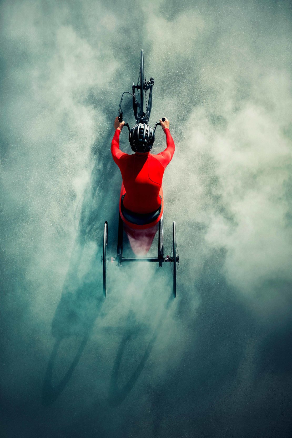 Overhead shot of an adult wearing red sportswear and a helmet riding a specialized handcycle, surrounded by a cloud of white powder.