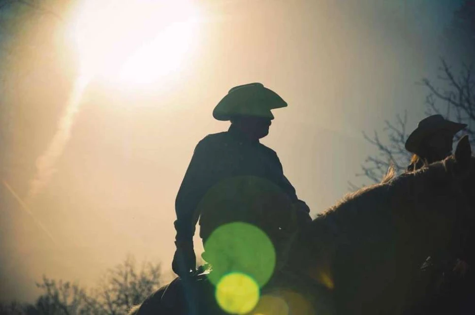 Two silhouetted cowboys on horseback under a bright sun, capturing a Western aesthetic in an outdoor rural environment.