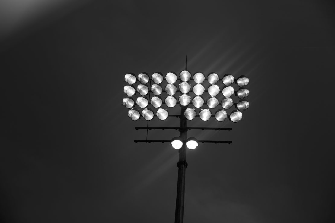 A black and white image of a stadium light array with multiple bulbs mounted on a tall metal pole against a dark sky background.