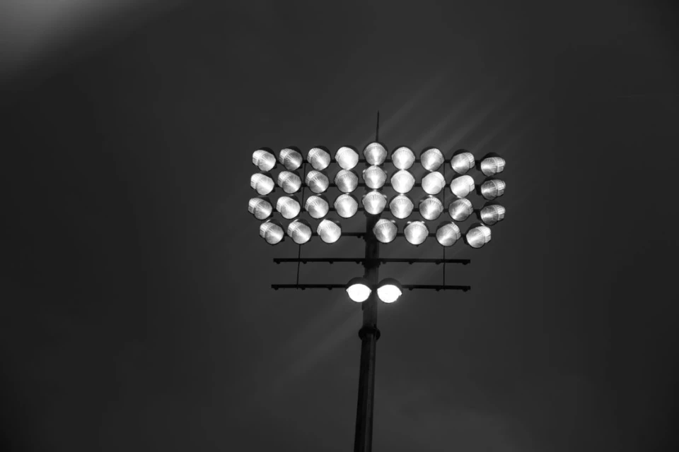 A black and white image of a stadium light array with multiple bulbs mounted on a tall metal pole against a dark sky background.