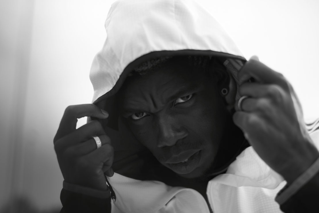 Monochrome portrait of a serious-looking adult male with dark skin, wearing a white hooded jacket, with his hands on the hood strings.