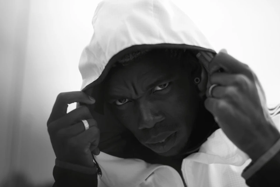 Monochrome portrait of a serious-looking adult male with dark skin, wearing a white hooded jacket, with his hands on the hood strings.