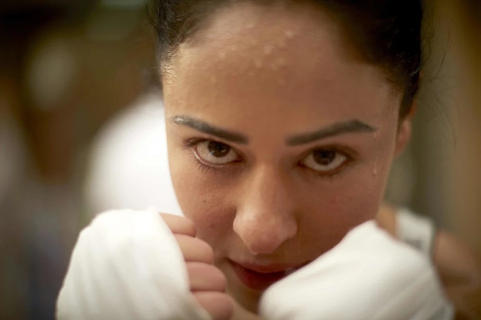 Close-up of a sweaty adult woman in boxing stance, with wrapped fists and intense gaze, set against a blurred indoor background.