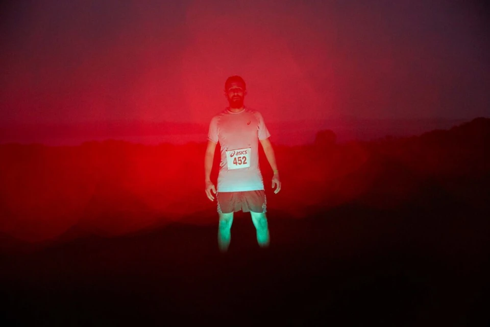 A man stands illuminated by a bright red and green light while wearing a race bib against a dark landscape background.