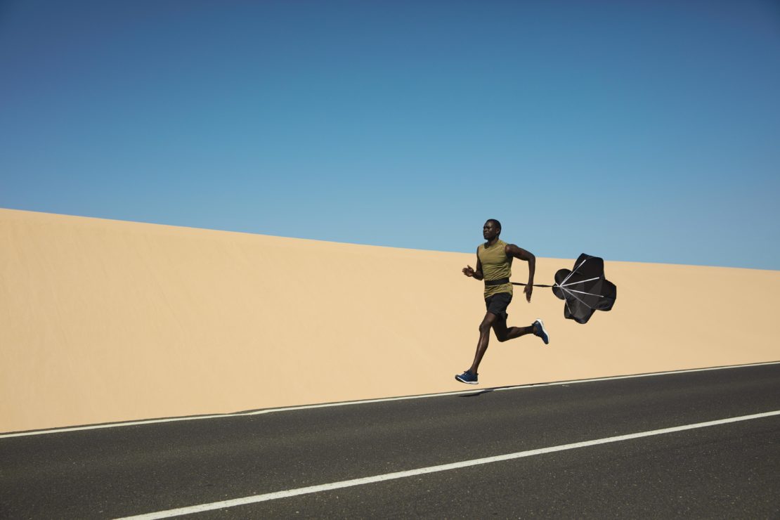An African American man runs on an asphalt road, pulling a black parachute-style training device, against a blue sky and neutral wall backdrop.