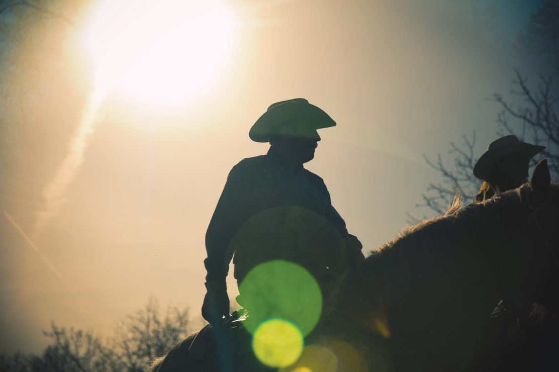 Silhouette of two cowboys on horseback against a bright sky, wearing hats, creating a dramatic and timeless Western scene.