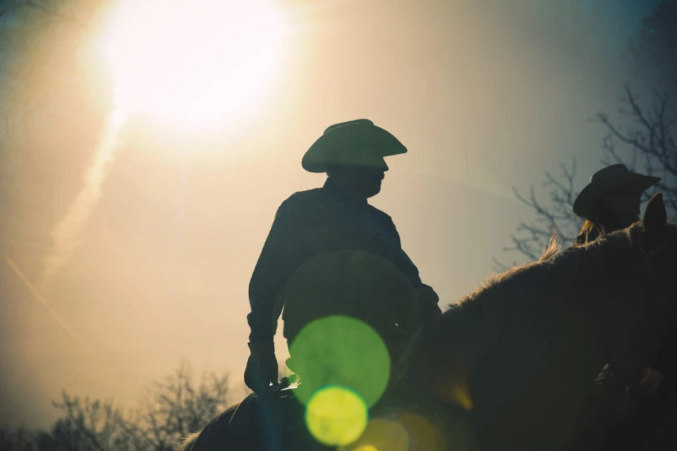 Silhouette of two cowboys on horseback against a bright sky, wearing hats, creating a dramatic and timeless Western scene.