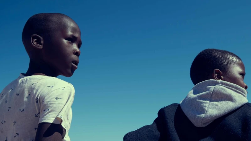 Two young African boys stand outdoors against a bright blue sky, looking off to the side. One wears a t-shirt with anchor patterns.