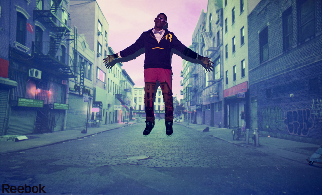 Man in streetwear posing in an urban setting with buildings and street visible. He is wearing Reebok apparel and sneakers.