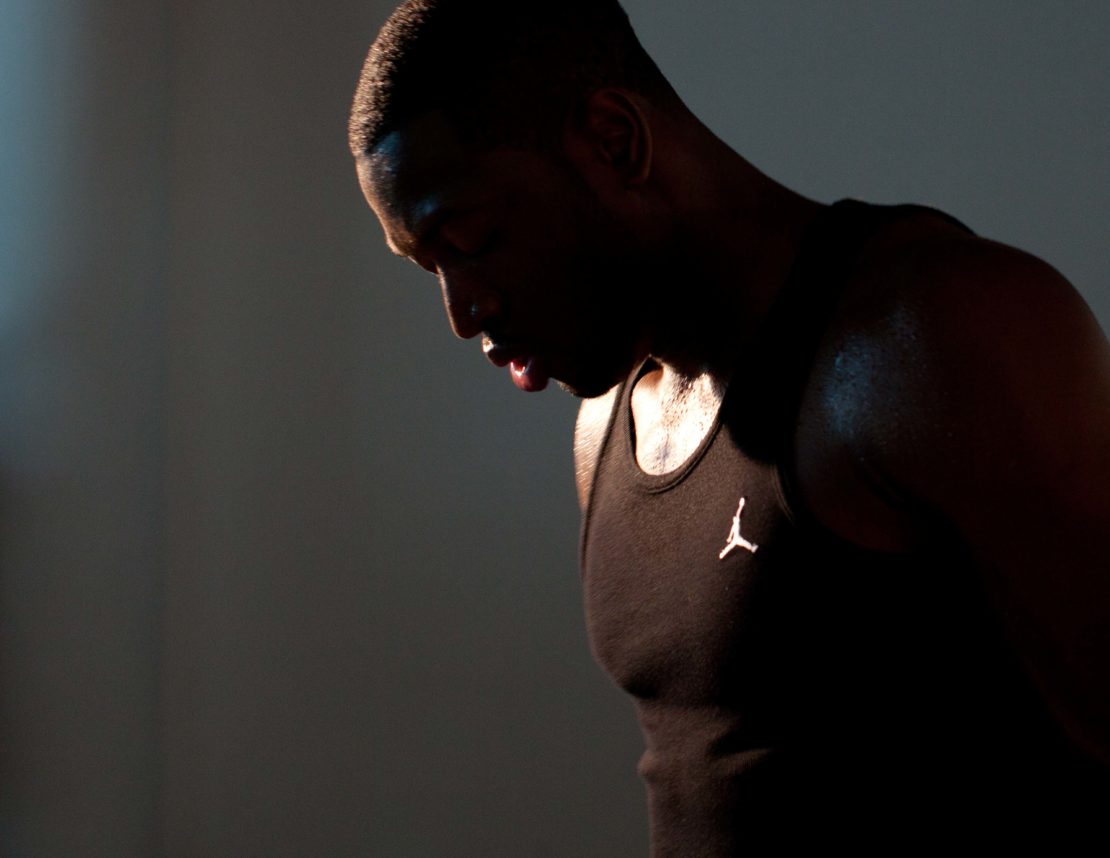 Man in black tank top with logo looking down in a dimly lit room with sweat on his shoulder and face. Studio shot.