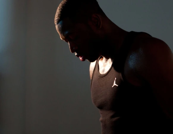 Man in black tank top with logo looking down in a dimly lit room with sweat on his shoulder and face. Studio shot.