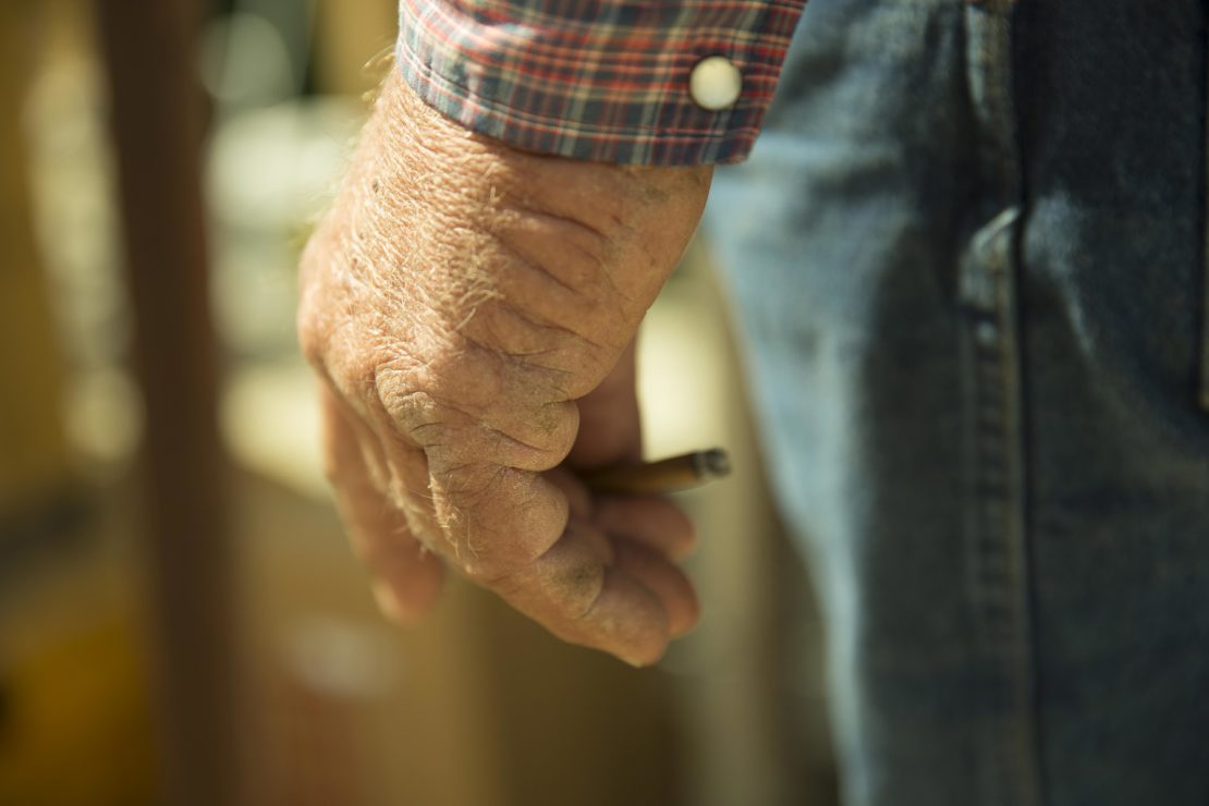 Close-up of an elderly man's weathered hand holding a cigarette, wearing a plaid shirt and blue jeans.