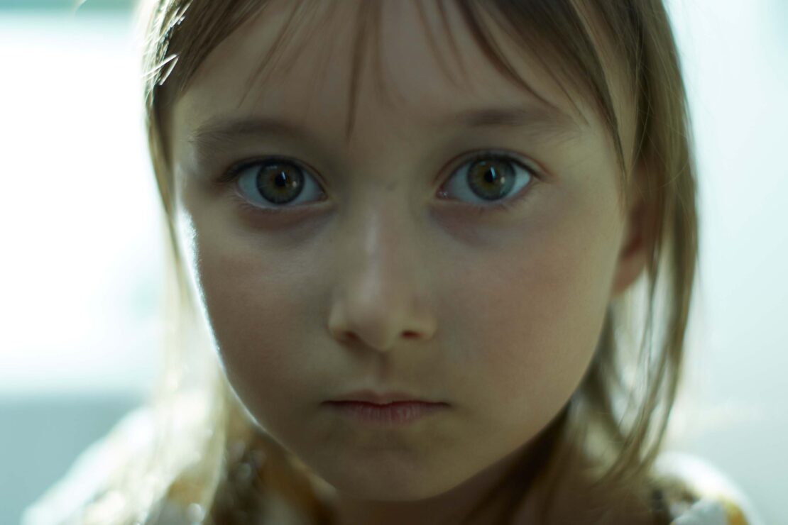Close-up portrait of a young girl with brown hair and captivating brown eyes, looking directly at the camera with a serious expression.