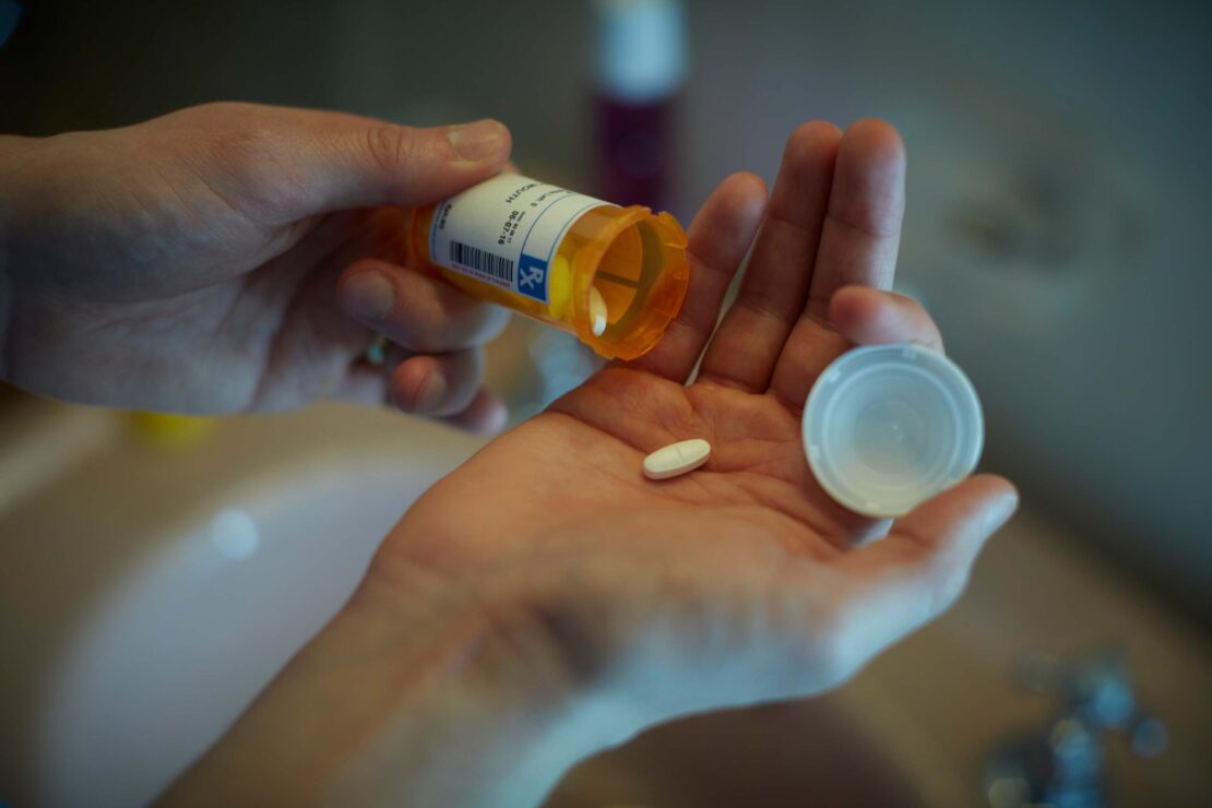 Close-up shot of a person pouring a white pill from a prescription bottle into their hand in a bathroom setting, healthcare.