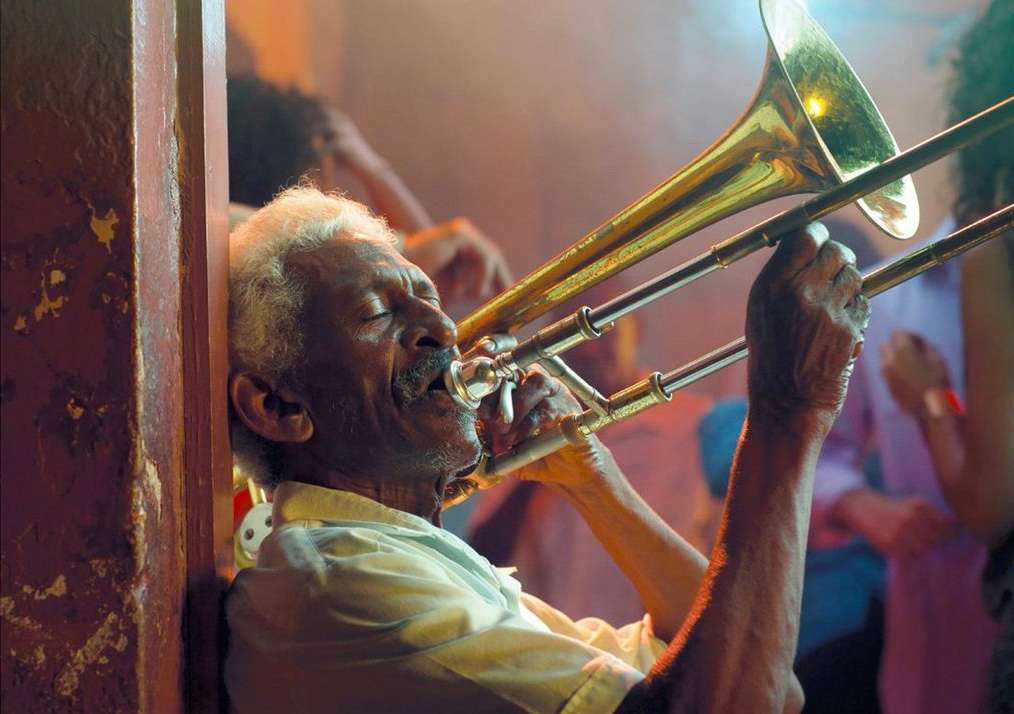 Older African-American musician playing trombone in an intimate performance space, with party-goers dimly visible in the background.