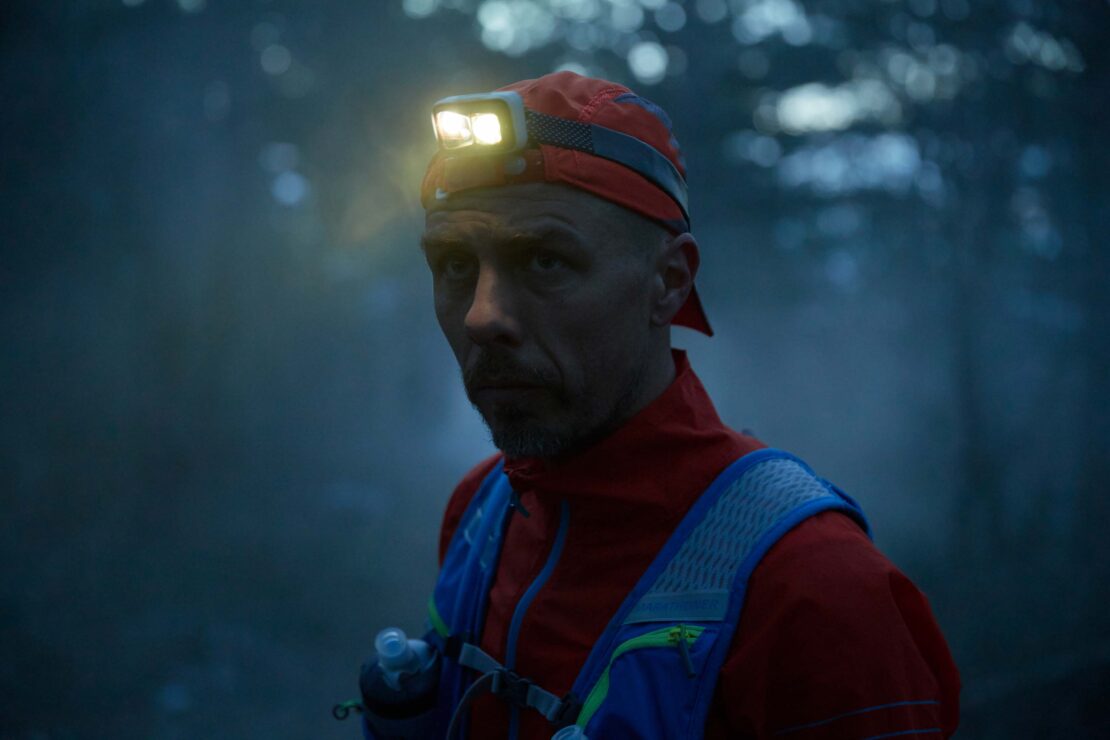 Man wearing headlamp and running vest in dark forest, looking at camera with serious expression. Outdoor fitness and adventure in nature.