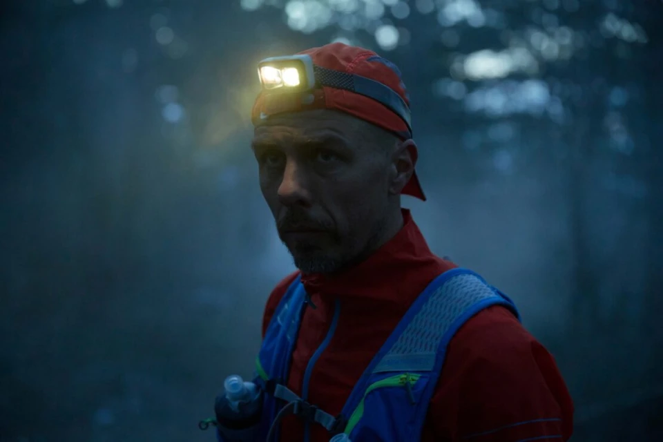 Man wearing headlamp and running vest in dark forest, looking at camera with serious expression. Outdoor fitness and adventure in nature.