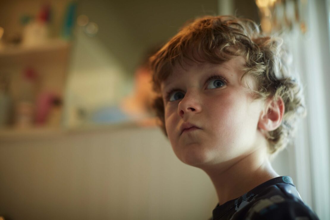 Close-up portrait of a young boy with curly hair looking up. Indoor setting with diffused natural light, shallow depth of field.