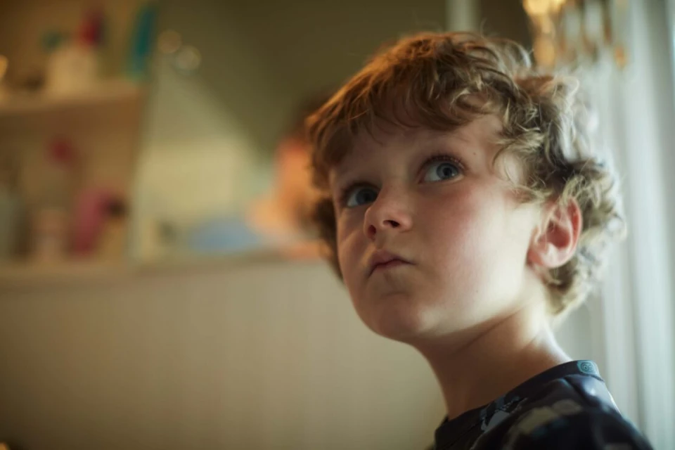 Close-up portrait of a young boy with curly hair looking up. Indoor setting with diffused natural light, shallow depth of field.
