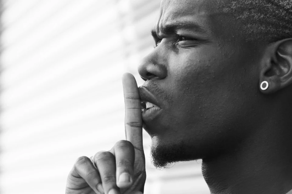 Black and white portrait of a dark-skinned adult male holding a finger to his lips, indicating silence, with an earring detail.
