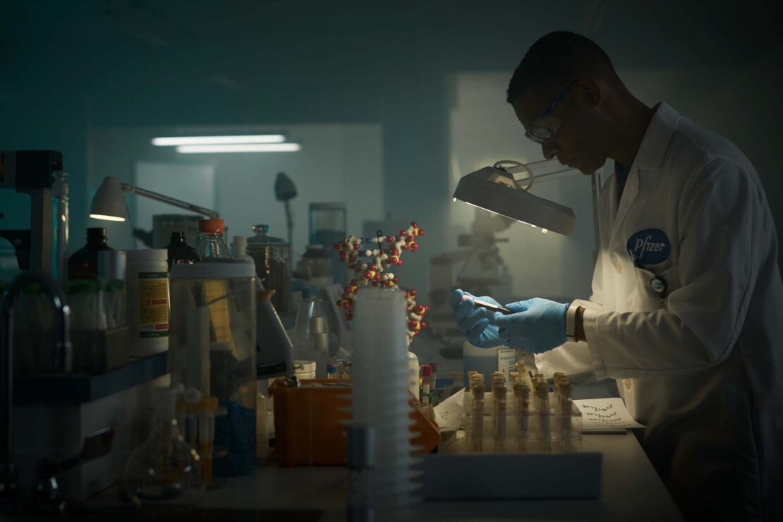 Scientist in lab coat and safety glasses examines sample in a laboratory setting with equipment, beakers, and model molecules. 