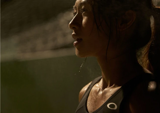 Profile view of an athletic adult woman with sweat on her face and chest. She's wearing a sports bra in a dark, hazy environment.