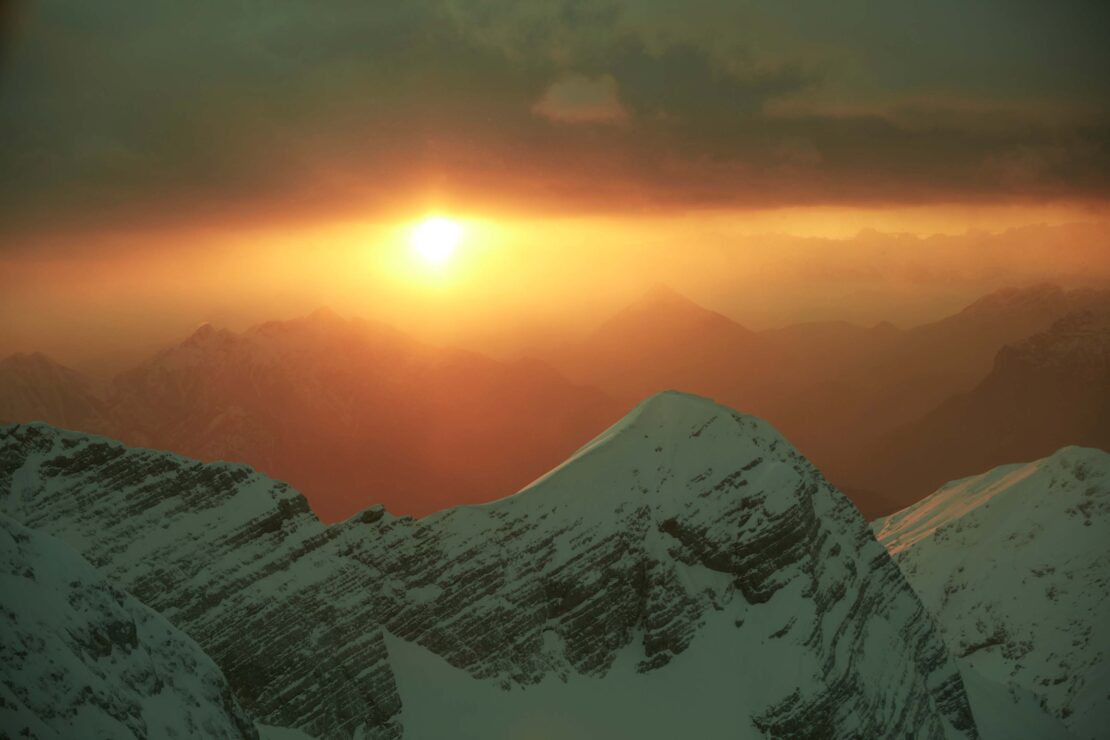 Scenic view of snow-capped mountains with the sun glowing brightly in the sky, creating a warm and ethereal atmosphere.