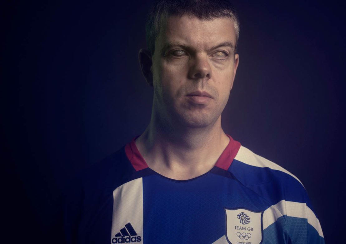 Portrait of a man wearing a blue and white Team GB sports shirt, set against a dark background with subtle lighting.