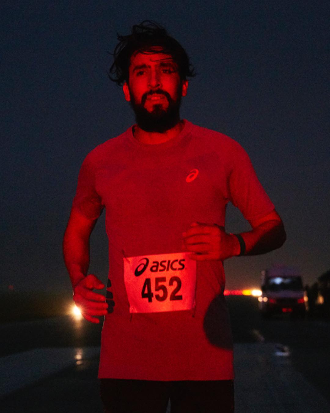 A runner with a beard and a bib participates in a road race at night. Headlights from vehicles are visible in the background.