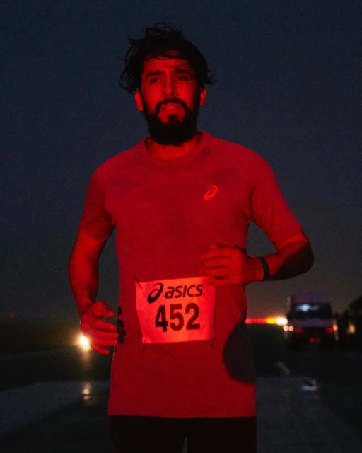 A runner with a beard and a bib participates in a road race at night. Headlights from vehicles are visible in the background.