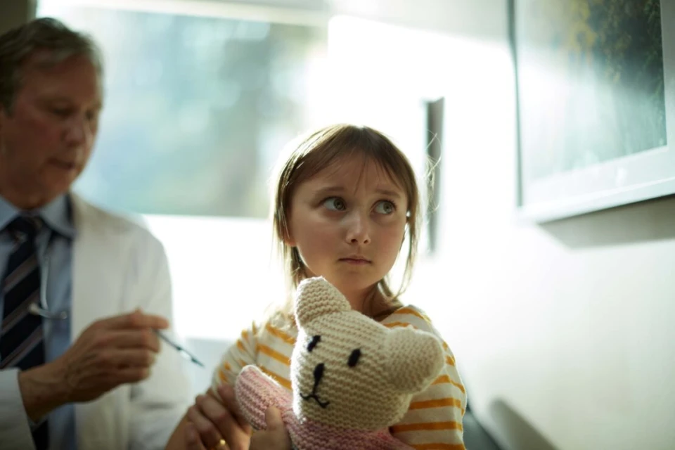 Doctor about to give a shot to a young girl holding a teddy bear in a medical office under bright, natural light, concerned facial expression.