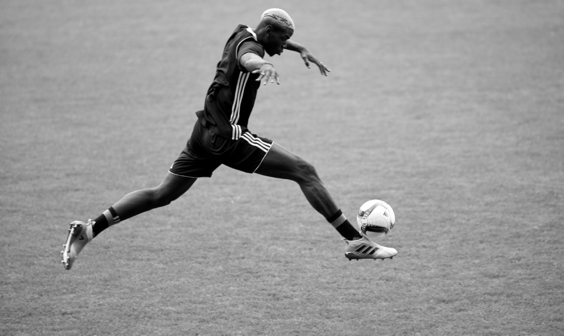 Black and white photo of a soccer player in mid-air kicking a soccer ball on a grass field during a soccer match, sport, action.