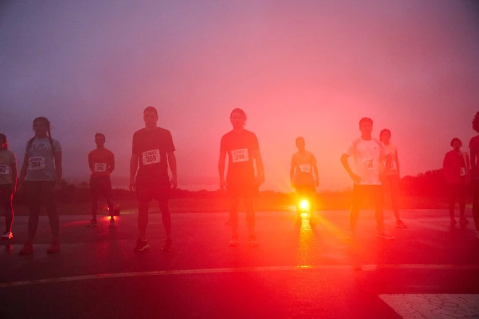 Group of runners standing on a road at twilight, wearing race bibs and athletic wear, illuminated by a red light source.