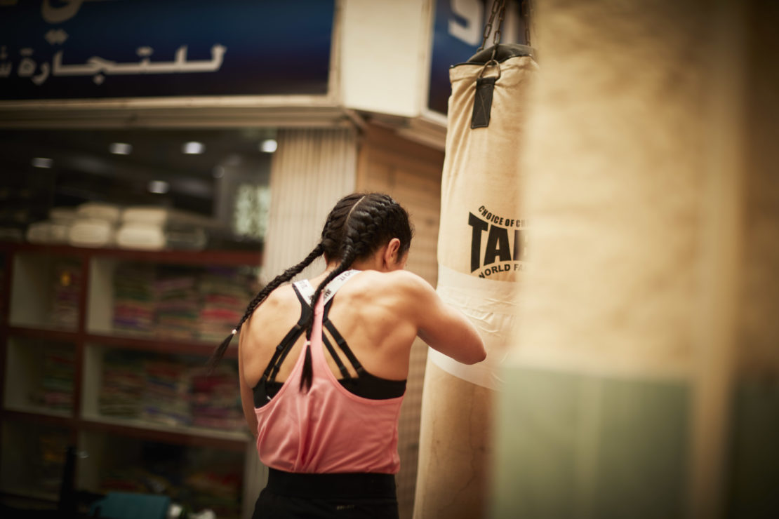 A muscular woman with braids punches a tan punching bag in a gym. The woman wears a pink tank top and black sports bra.
