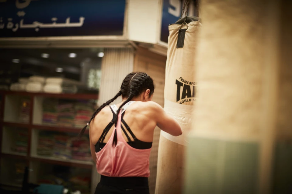 A muscular woman with braids punches a tan punching bag in a gym. The woman wears a pink tank top and black sports bra.
