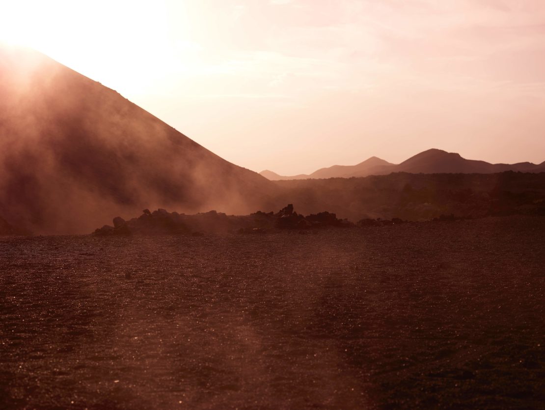 Volcanic landscape with mountains and dusty ground, illuminated by soft sunlight creating a serene, natural scene in a desolate setting.
