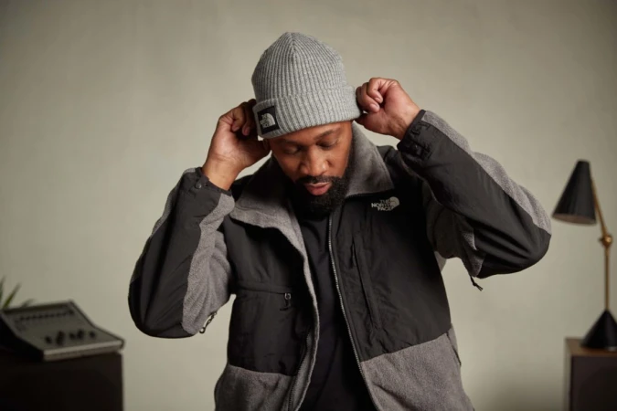 Man wearing a grey beanie and fleece jacket adjusting his hat against a neutral studio background