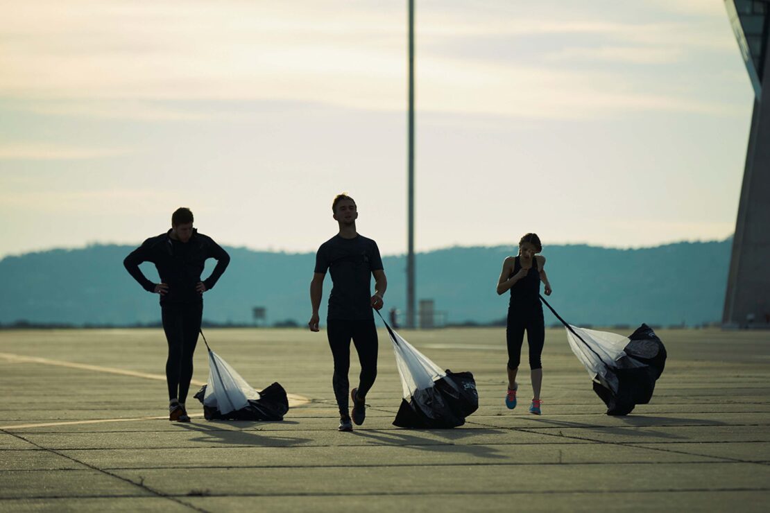 Three athletes in activewear are pulling resistance parachutes on a tarmac runway with an airplane hangar in the background.