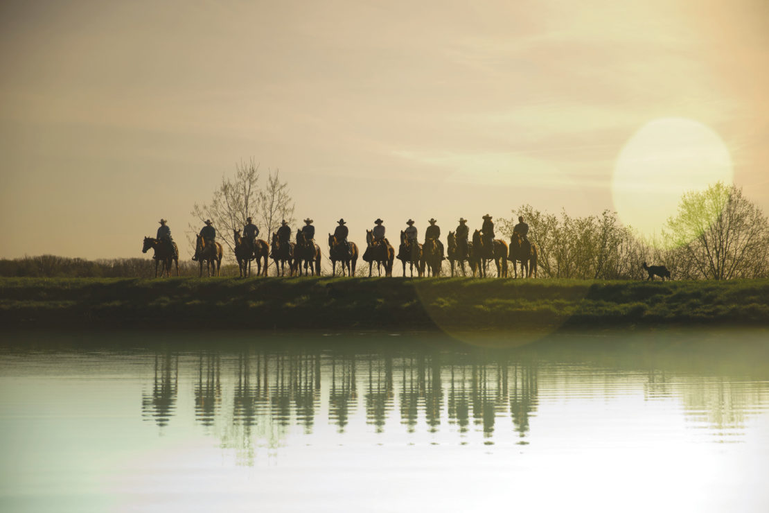 Silhouetted group of cowboys riding horses along a grassy ridge reflected in water at golden hour with a dog running nearby.