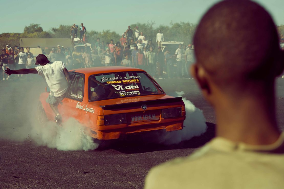 Orange sedan drifting on asphalt with a man hanging off the side door, surrounded by white smoke, and a crowd in the background.